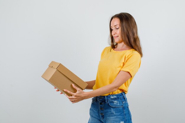 Jonge vrouw in t-shirt, korte broek met kartonnen doos en glimlachen.