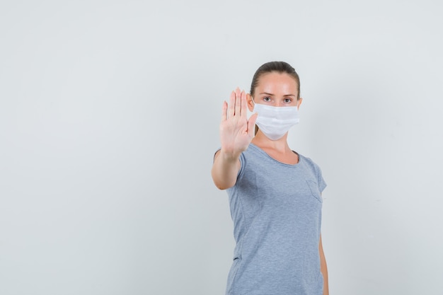 Jonge vrouw in t-shirt, masker dat weigeringsgebaar toont en ernstig, vooraanzicht kijkt.