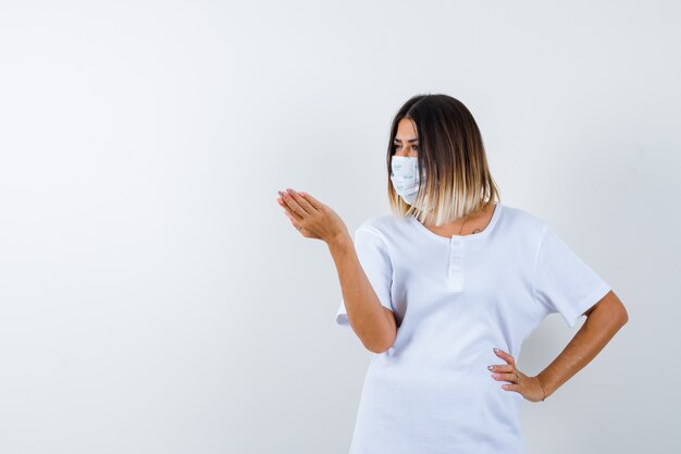 Jonge vrouw in t-shirt, masker strekken hand in vragend gebaar terwijl hand op taille en op zoek ernstig, vooraanzicht.