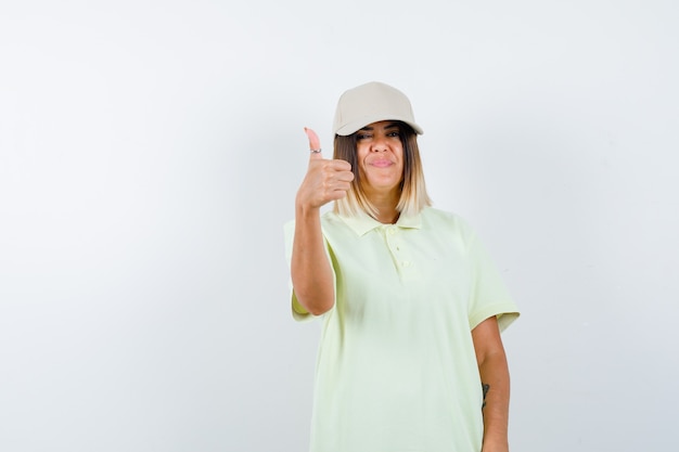 Jonge vrouw in t-shirt, pet met duim omhoog en op zoek vrolijk, vooraanzicht.