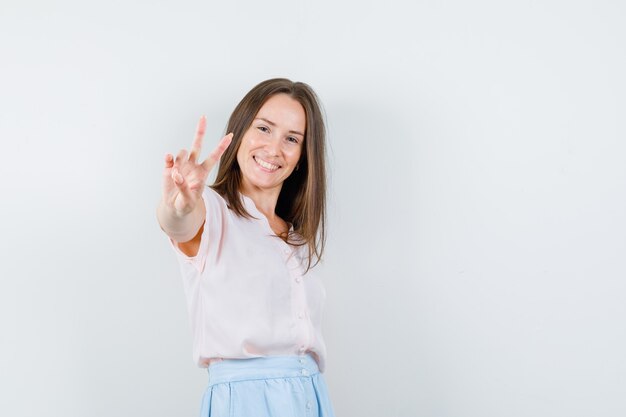 Jonge vrouw in t-shirt, rok die overwinningsteken toont en positief, vooraanzicht kijkt.