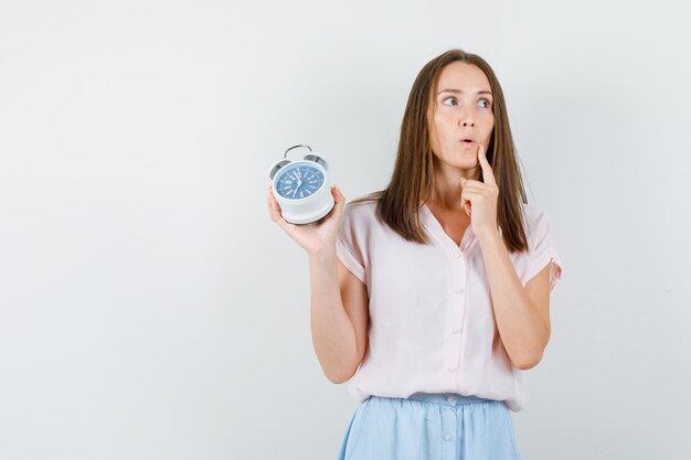 Jonge vrouw in t-shirt, rok die wekker houdt en peinzend kijkt, vooraanzicht.