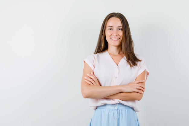 Jonge vrouw in t-shirt, rok die zich met gekruiste armen bevindt en positief, vooraanzicht kijkt.