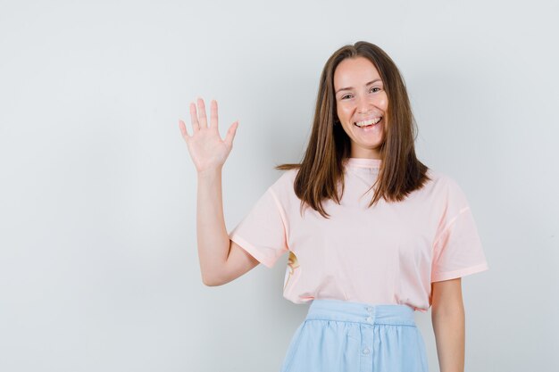 Jonge vrouw in t-shirt, rok zwaaiende hand voor groet en op zoek gelukkig, vooraanzicht.