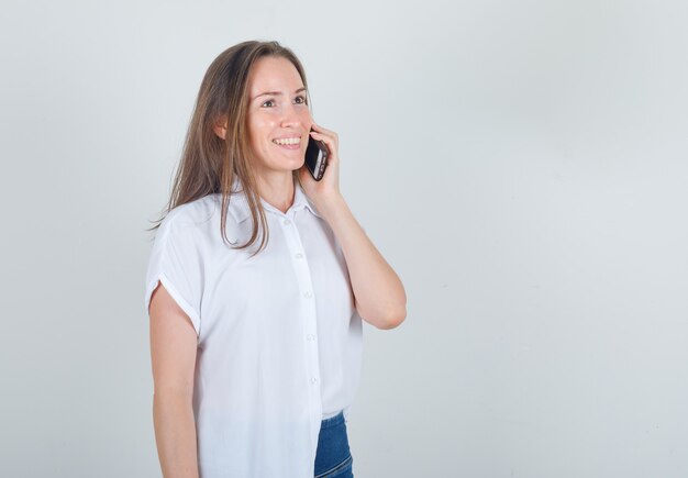 Jonge vrouw in t-shirt, spijkerbroek praten over smartphone en op zoek vrolijk