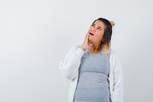 Jonge vrouw in t-shirt, vest die hand op de wang houdt en peinzend kijkt, vooraanzicht.