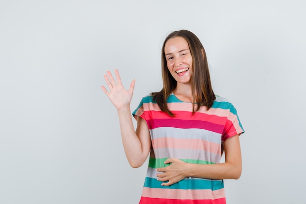 Jonge vrouw in t-shirt zwaaiende hand en knipogen oog, vooraanzicht.