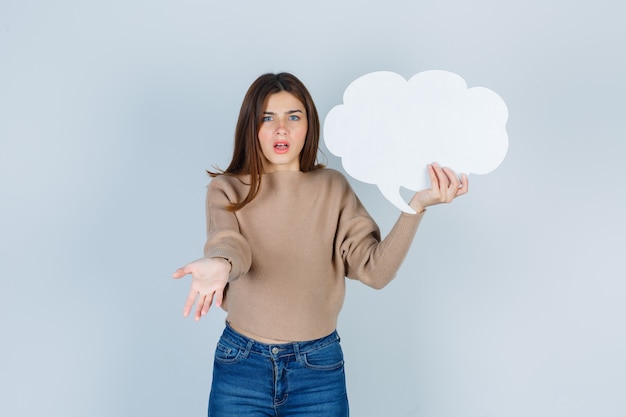 Jonge vrouw in trui, jeans die ontevreden is over domme vraag, papieren tekstballon houdt en er somber uitziet, vooraanzicht.