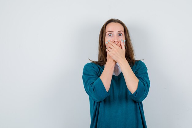 Jonge vrouw in trui over shirt met handen op mond en geschokt, vooraanzicht.