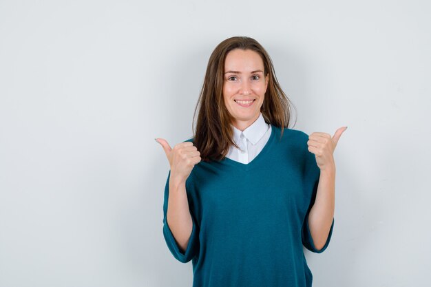 Jonge vrouw in trui over shirt wijst naar de tegenovergestelde richtingen met duimen en ziet er vrolijk uit, vooraanzicht.