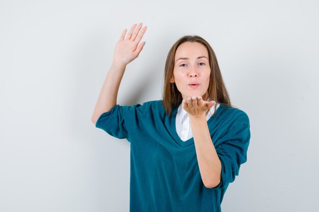 Jonge vrouw in trui over wit overhemd blaast luchtkus met pruilende lippen, steekt open hand op en ziet er vredig uit, vooraanzicht.