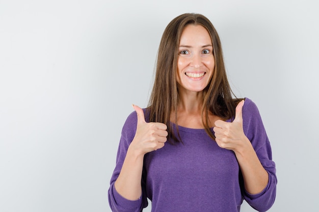 Gratis foto jonge vrouw in violet overhemd duimen opdagen en op zoek vrolijk, vooraanzicht.