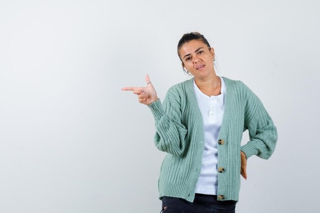 Jonge vrouw in wit overhemd en mintgroen vest wijzend naar links met wijsvinger terwijl ze de hand op de taille houdt en er serieus uitziet