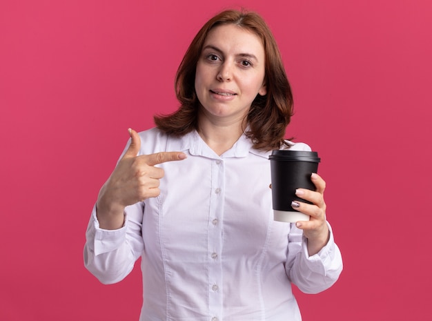 Jonge vrouw in wit overhemd met koffiekopje wijzend met wijsvinger naar het glimlachend zelfverzekerd staande over roze muur