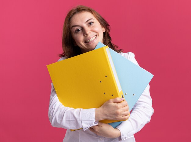 Jonge vrouw in wit overhemd met mappen kijken voorkant lachend met blij gezicht staande over roze muur