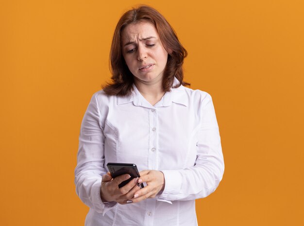 Jonge vrouw in wit overhemd met smartphone die ernaar kijkt met droevige uitdrukking die zich over oranje muur bevindt