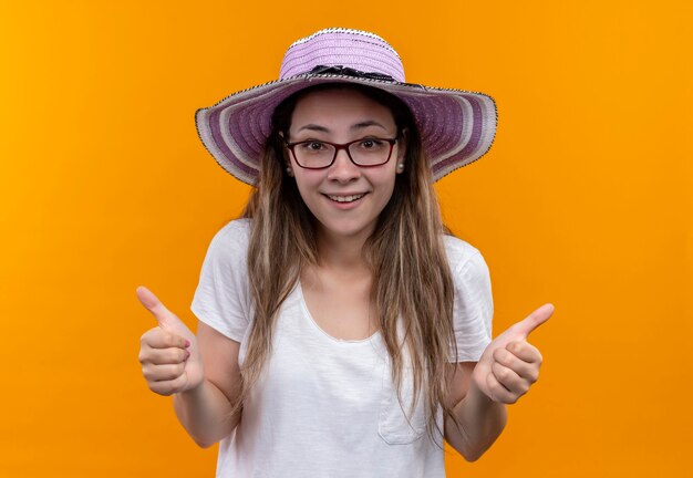 Jonge vrouw in wit t-shirt die de zomerhoed draagt die vrolijk glimlachend duimen toont die zich over oranje muur bevinden