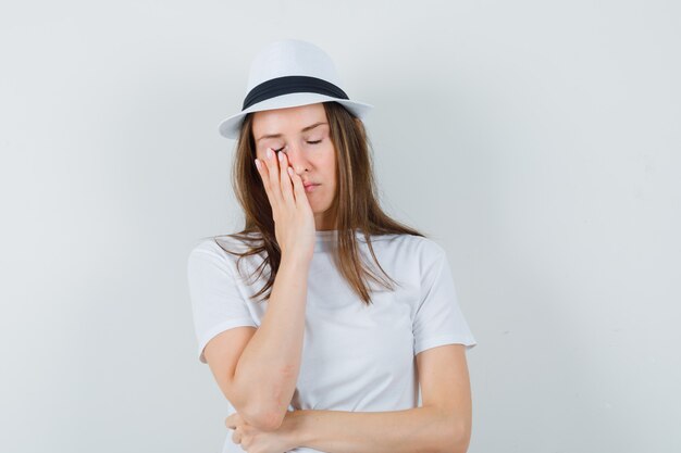 Jonge vrouw in wit t-shirt, hoed die hand op gezicht houdt en vermoeid kijkt.