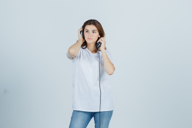 Jonge vrouw in wit t-shirt, spijkerbroek koptelefoon opstijgen en peinzend, vooraanzicht kijken.