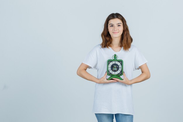 Jonge vrouw in wit t-shirt, spijkerbroek met klok en op zoek vrolijk, vooraanzicht.