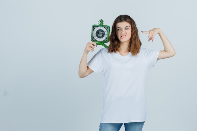 Jonge vrouw in wit t-shirt, spijkerbroek wijzend op de klok, fronsend gezicht en op zoek verontrust, vooraanzicht.