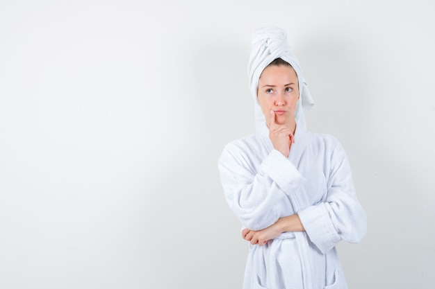 Jonge vrouw in witte badjas, handdoek die vinger dichtbij mond houdt en peinzend, vooraanzicht kijkt.