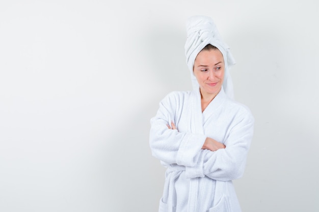 Jonge vrouw in witte badjas, handdoek die zich met gekruiste armen bevindt en peinzend, vooraanzicht kijkt.