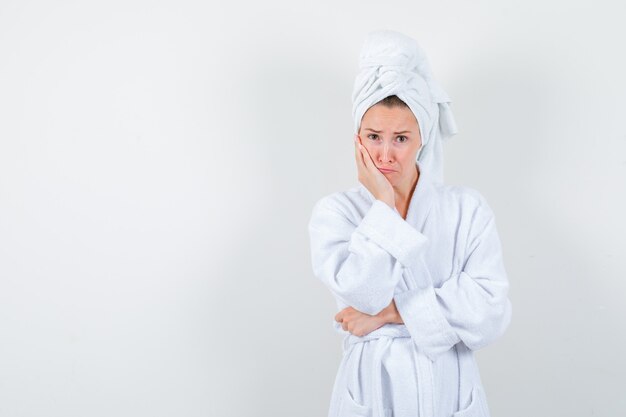 Jonge vrouw in witte badjas, handdoek leunende wang op opgeheven handpalm en op zoek verdrietig, vooraanzicht.