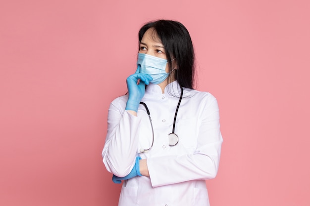 jonge vrouw in witte medische pak blauwe handschoenen blauw beschermend masker met stethoscoop op roze