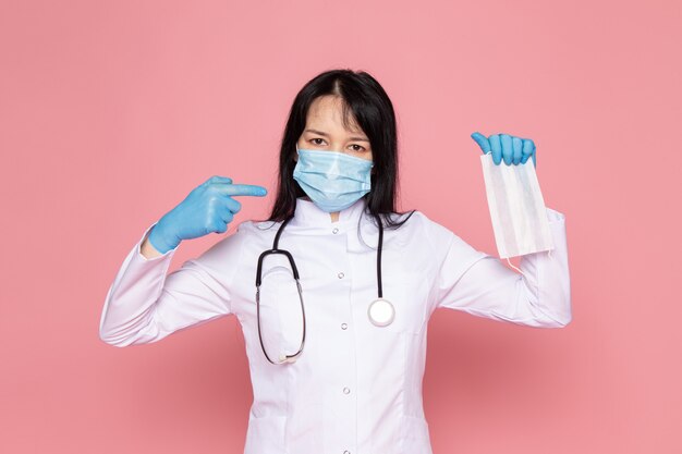 jonge vrouw in witte medische pak blauwe handschoenen blauw beschermend masker met stethoscoop op roze