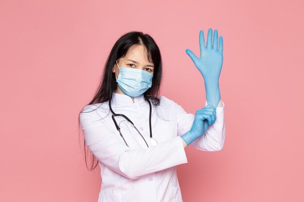 jonge vrouw in witte medische pak blauwe handschoenen blauw beschermend masker met stethoscoop op roze