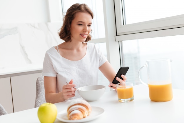Jonge vrouw in witte t-shirt met behulp van mobiele telefoon tijdens het ontbijt in de keuken
