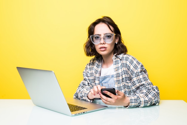 Jonge vrouw in zonnebril texting zittend op haar bureau op haar mobiele telefoon met laptop vooraan