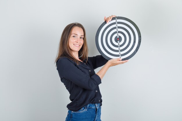 Jonge vrouw in zwart overhemd, jeansborrels die dartbord houden en vrolijk kijken