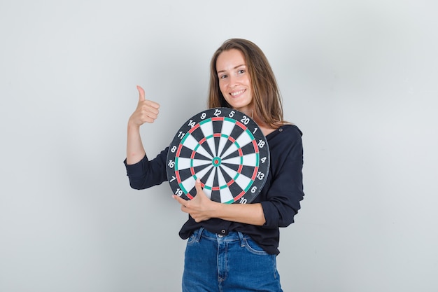 Jonge vrouw in zwart shirt, jeans broek dartbord met duim omhoog houden en glimlachen