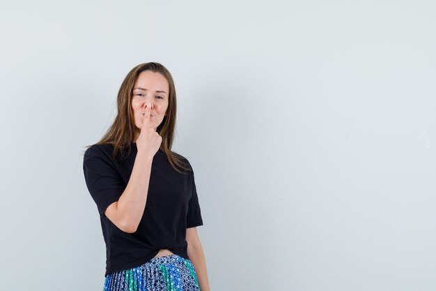 Jonge vrouw in zwart t-shirt en blauwe rok wijsvinger op mond zetten en gelukkig glimlachen en kijken