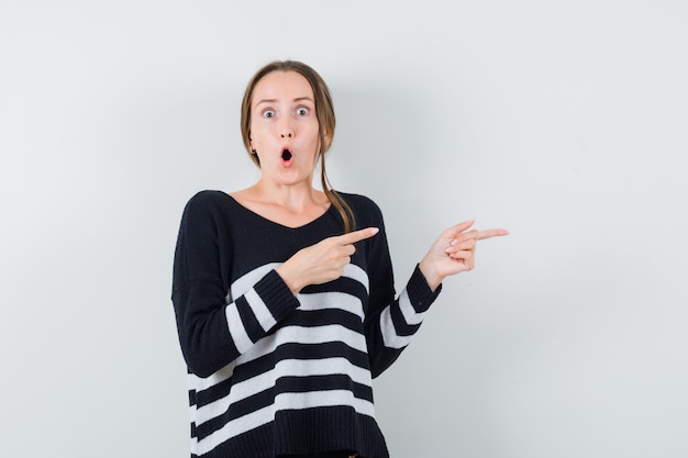 Jonge vrouw in zwarte blouse en zwarte broek die met wijsvingers naar rechts wijst en verbaasd kijkt