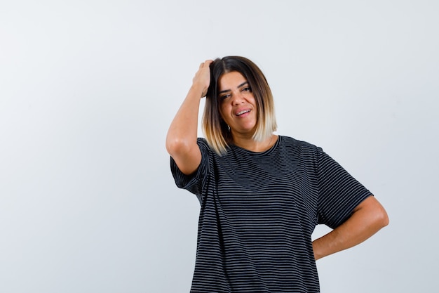 Jonge vrouw in zwarte jurk die een hand op het hoofd houdt, een andere hand op de taille en er mooi uitziet, vooraanzicht.