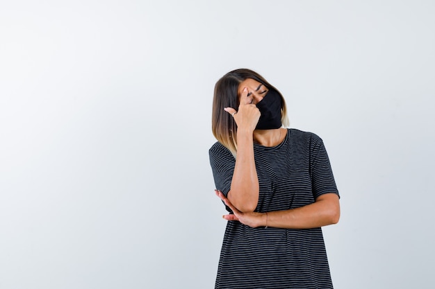 Jonge vrouw in zwarte jurk, zwart masker wijsvinger op voorhoofd te zetten, een hand onder de elleboog te houden en uitgeput, vooraanzicht te kijken.