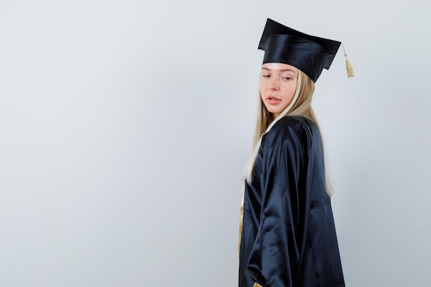 Gratis foto jonge vrouw kijkt over haar schouder in afgestudeerd uniform en ziet er aantrekkelijk uit. .