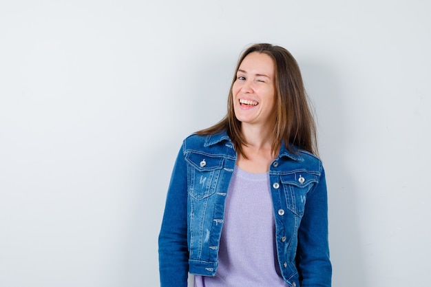 Jonge vrouw knipoogt terwijl ze in t-shirt, spijkerjasje poseert en er vrolijk uitziet. vooraanzicht.