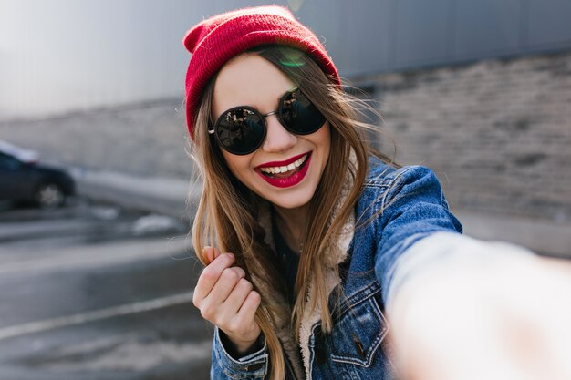 Jonge vrouw lachen in casual rode hoed selfie maken. Openluchtportret van prachtig meisje die zich voordeed op stedelijke muur met oprechte glimlach.