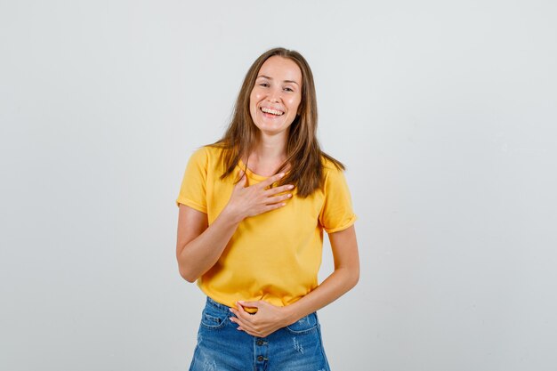 Jonge vrouw lachen met handen op de borst en buik in t-shirt, korte broek en op zoek gelukkig. vooraanzicht.