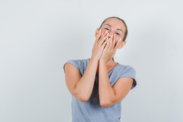 Jonge vrouw lachen met handen op mond in grijs t-shirt vooraanzicht.