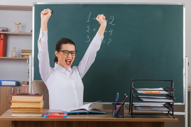 Jonge vrouw leraar bril met boek uitleggen les op zoek gek gelukkig gebalde vuisten zittend op school bureau voor schoolbord in klas