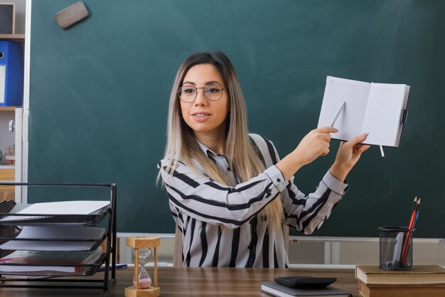 jonge vrouw, leraar, vervelend, bril, zittende, op, school, bureau, voor, blackboard, in, klaslokaal, controleren, huiswerk, van, studenten, uitleggen, les, kijkend, zelfverzekerd