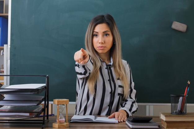 jonge vrouw leraar zit op school bureau voor schoolbord in de klas huiswerk controleren van studenten wijzen met wijsvinger naar camera kijken zelfverzekerd