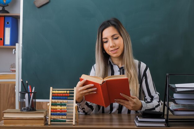 jonge vrouw leraar zit op school bureau voor schoolbord in klas leesboek voorbereiden op les glimlachend zelfverzekerd