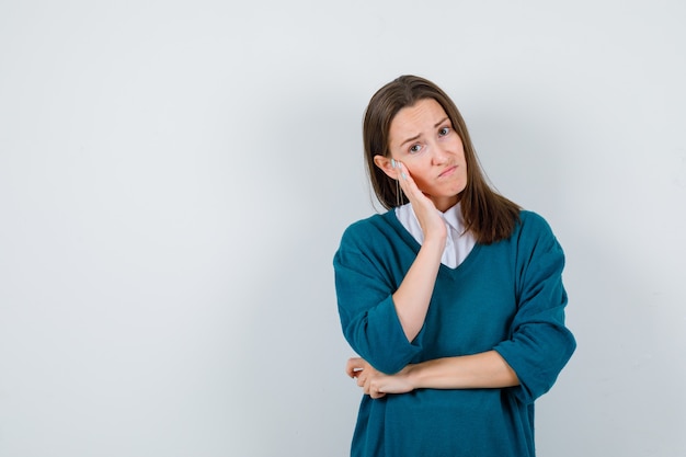 Jonge vrouw leunt wang op hand in trui over wit overhemd en kijkt teleurgesteld, vooraanzicht.