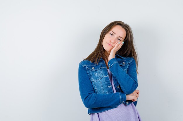 Jonge vrouw leunt wang op palm in t-shirt, jas en ziet er moe uit, vooraanzicht.